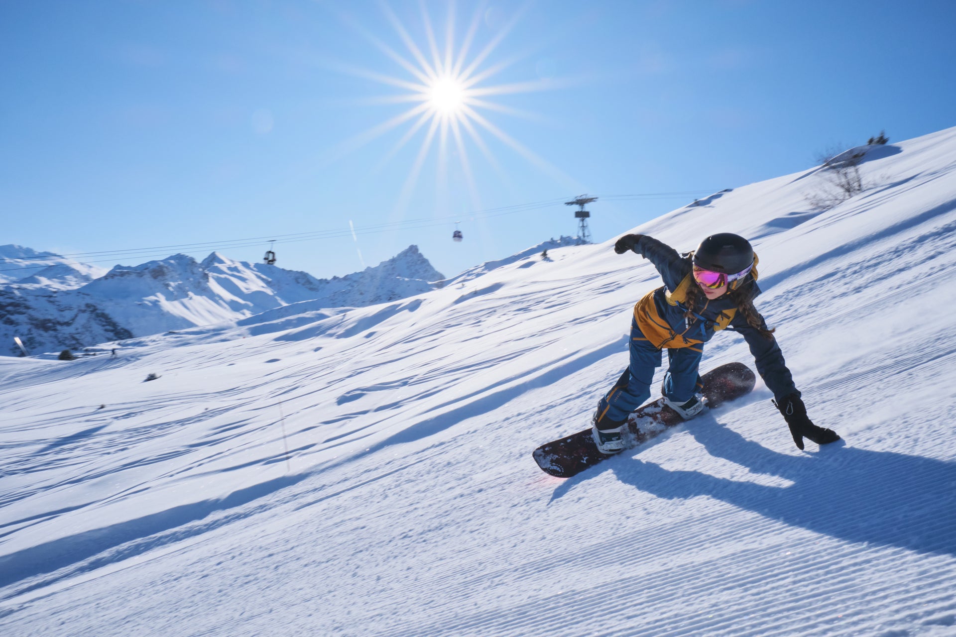 Untrakt | A snowboarder in a blue and yellow suit carving on a sunny, snow-covered slope with ski lifts in the background